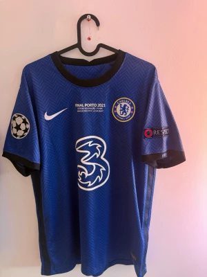 Chelsea matchtröja Kanté 2021 - Blå Chelsea matchtröja från Champions League-finalen 2021 med Kanté och nummer 7 på ryggen. Tröjan har svarta detaljer, officiella loggor och tryck, samt Champions League-märke på ärmen. Tillverkad i lätt och ventilerande material.