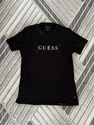 Guess t-shirt - Snygg svart t-shirt från Guess med stor broderad logga på bröstet. Slim fit-modell i 100% ekologisk bomull. Klassisk rund halsringning och diskreta sömdetaljer runt loggan för en cool touch.