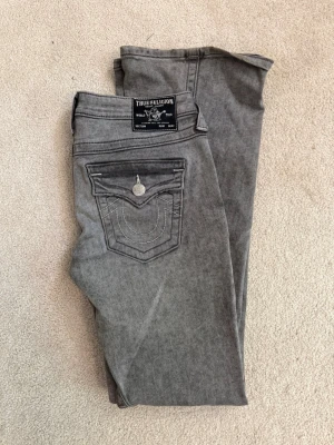 Grå bootcut jeans True Religion - Snygga grå jeans från True Religion med bootcut passform och klassiska kontrastsömmar. Jeansen har fem fickor, silverfärgade detaljer och ikonisk fickdesign bak. Perfekta för dig som vill ha en cool och avslappnad stil. Storleken motsvarar s/m