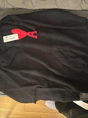 Svart stickad tröja med röd logga - Snygg svart stickad tröja med stor röd hjärtformad logga och bokstaven A på bröstet. Tröjan har rund halsringning och ribbade muddar vid ärmslut och nederkant. Perfekt för dig som gillar stilrena plagg med en twist.