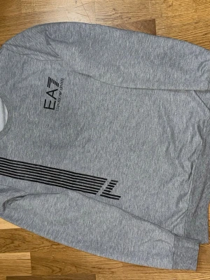 Grå sweatshirt från Emporio Armani - Snygg grå sweatshirt från Emporio Armani EA7 med svart tryck på bröstet och coolt linjemönster längs sidan. Tröjan har rund halsringning och ribbade muddar vid ärmslut och nederkant. Perfekt för en avslappnad och trendig look.