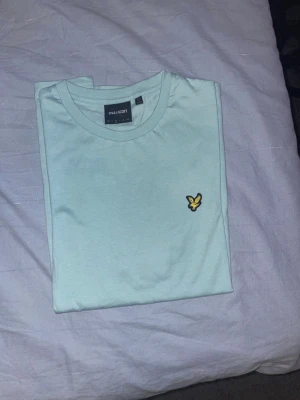Ljusblå t-shirt från Lyle & Scott - Snygg ljusblå t-shirt från Lyle & Scott med klassisk gul logga på bröstet. T-shirten har rund halsringning och korta ärmar. Tillverkad i mjuk bomull som känns skön mot huden. Perfekt för en clean och enkel stil.