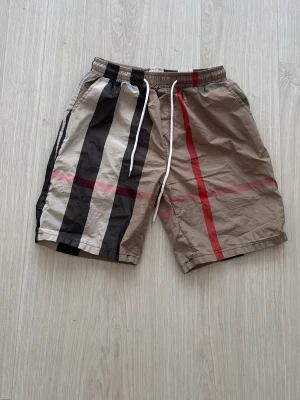 Burrbery shorts - Snygga shorts i beige med breda svarta, vita och röda ränder. De har elastisk midja med vit snörning för skön passform. Materialet känns lätt och luftigt, perfekt för varma dagar. Stilen är avslappnad och mönstret ger en trendig vibe.