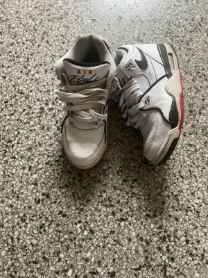 Nike Air Flight vita sneakers - Nike Air Flight sneakers i vitt läder med svarta och röda detaljer. Klassisk retrodesign med tydlig Nike-logga på sidan och röd logga på sulan. Skorna har tjock sula och vadderad plös för extra komfort. Perfekta för dig som gillar old school streetstyle.
