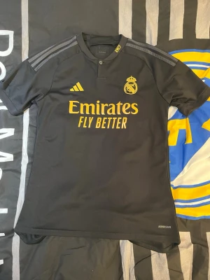 Äkta Real Madrid tröja - Real Madrid 23/24 tredjeställ. från Adidas med gula detaljer och klubbmärke på bröstet. Tröjan har korta ärmar, knapp i halsen och 'Emirates Fly Better' tryckt framtill. Tillverkad i lätt och ventilerande funktionsmaterial.