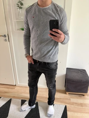  Hollister - Snygg grå stickad tröja från Hollister med rund halsringning och diskret logga på bröstet. Tröjan har långa ärmar och ribbade muddar vid ärmslut och nederkant. Perfekt till vardags för en avslappnad men stilren look.