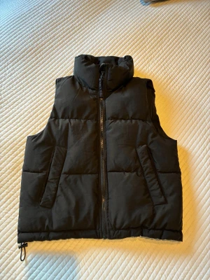 Svart väst med hög krage storlek 140/152. - Snygg svart väst med hög krage och dragkedja framtill. Västen har två snedställda fickor och justerbar dragsko nedtill. Perfekt för lager på lager och ger en clean look till din outfit.