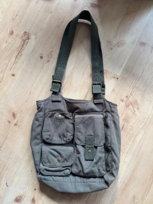 Grön axelväska med fickor - Säljer en grön väska i canvas med flera praktiska ytterfickor framtill och en ficka bak. Väskan har justerbara axelband i grönt tyg och en rymlig insida. Perfekt för dig som behöver en ny skolväska till exempel ! Budas ut på min live 25 augusti 19.00. 