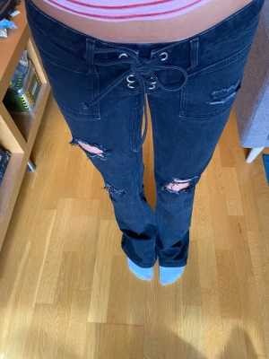 Svarta slitna jeans med snörning - Säljer ett par svarta jeans med snörning i midjan och flera slitningar på benen. Jeansen har raka ben och stora fickor framtill. Perfekta för en edgy och avslappnad stil.