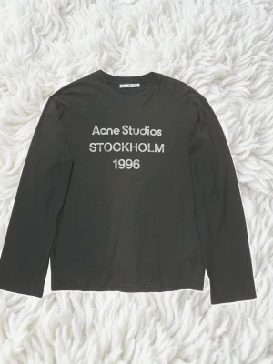 Svart långärmad topp Acne Studios - Svart långärmad topp från Acne Studios med tryck 'Acne Studios STOCKHOLM 1996' på bröstet. Tillverkad i 100% bomull för en skön och avslappnad känsla. Perfekt till jeans eller kjol för en clean och stilren look.