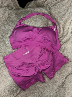 Träningsset - Säljer ett matchande set med sport-bh och shorts i en snygg rosa färg. Materialet är stretchigt och följsamt, perfekt för träning eller gym. Både bh:n och shortsen har logga framtill och ger en sportig look.