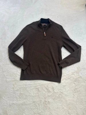 Brun halvzip tröja från Morris  - Snygg brun tröja med halvzip och hög krage. Tröjan har långa ärmar, ribbade muddar och en liten broderad logga på bröstet. Perfekt för lager på lager och enkel att matcha med jeans eller chinos.