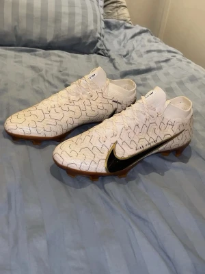 Vita Nike fotbollsskor med svart swoosh - Säljer ett par vita Nike fotbollsskor med svart swoosh och unikt mönster över hela skon. Skorna har högt skaft och snörning samt guldfärgad yttersula med Zoom-dämpning. Perfekta för dig som vill sticka ut på planen.