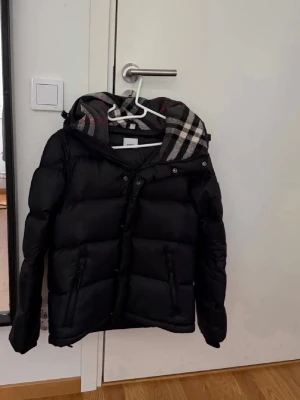 Svart Burberry jacka  - Snygg svart dunjacka med rutigt foder i huvan och dragkedja framtill. Jackan har två fickor med dragkedja och en avtagbar huva. Puffig modell som håller dig varm under kalla dagar. Kan även göras till väst till höst och vår!
