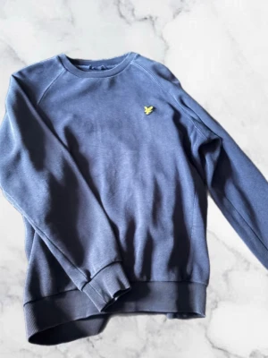 Lyle and scott tröja - Snygg mörkblå sweatshirt med rund hals och ribbade muddar. På bröstet sitter en liten gul broderad logga som ger en clean och stilren look. Perfekt till jeans eller joggers för en avslappnad stil.