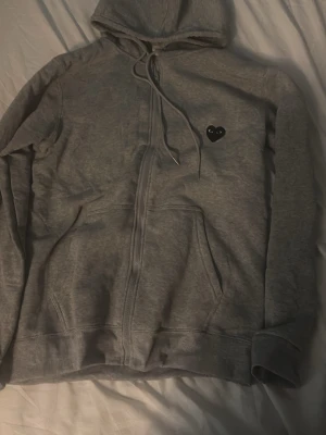 Grå Comme des garcons hoodie  - Säljer en grå zip-hoodie med huva. Hoodien har fickor framtill och ribbade muddar vid ärmar och nederkant. Perfekt för en chill och avslappnad stil.