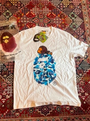 Vit BAPE t-shirt med blått camomönster - Säljer en vit t-shirt från BAPE med det klassiska apansikte-trycket i blått camomönster på bröstet. T-shirten har korta ärmar och rund hals. Perfekt för dig som gillar streetwear och vill sticka ut med en ikonisk design.