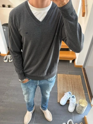 Grå V-ringad stickad tröja  - Snygg grå stickad tröja med V-ringning och diskret broderad logga på bröstet. Tröjan har långa ärmar och ribbade muddar vid ärmslut och nederkant. Perfekt att bära över en t-shirt för en clean och stilren look.