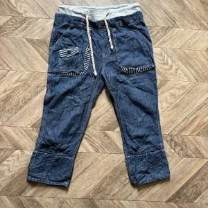 Blå jeans med dragsko och fickor - Säljer ett par blå jeans med grå mudd och vit dragsko i midjan. Jeansen har stora fickor fram och bak med randiga detaljer och knappstängning. Avslappnad passform och croppad längd, perfekta för en chill och casual stil.