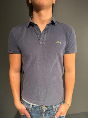 Mörkblå pikétröja från Lacoste - Klassisk mörkblå pikétröja från Lacoste med korta ärmar och krage. Tröjan har två knappar vid halsen och den ikoniska gröna krokodilloggan på bröstet. Perfekt till jeans eller shorts för en clean och stilren look. Modellen är 175cm 70 kg 
