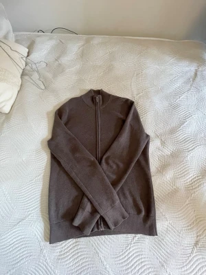 Brun stickad ziptröja från Bläck - Mysig brun stickad tröja från Bläck med hel dragkedja och hög krage. Perfekt för lager-på-lager och enkel att matcha med jeans eller chinos. Tröjan har en klassisk passform och ribbade muddar vid ärmslut och nederkant.