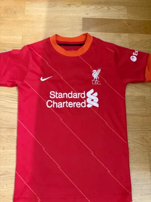 Liverpool matchtröja Salah #11 - Röd Liverpool matchtröja med vita detaljer, tryck av M. Salah och nummer 11 på ryggen. Nike-logga och klubbmärke på bröstet, sponsortryck på framsidan och Premier League-märke på ärmen. Orange ribbad krage och ärmslut. Perfekt för fotbollsfans!