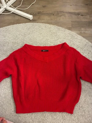 Röd stickad tröja från Gina Tricot - Snygg röd stickad tröja från Gina Tricot med bred v-ringning och ribbade muddar. Tröjan har en croppad passform och är gjord i ett mjukt, tjockt material som passar perfekt till höst och vinter. Enkel att styla med jeans eller kjol.