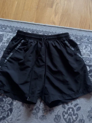 Svarta shorts från Calvin Klein - Snygga svarta shorts från Calvin Klein med logga längs sidorna och justerbar snörning i midjan. Perfekta för en avslappnad och sportig stil. Tillverkade i ett lätt och bekvämt material.