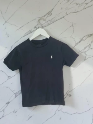 Ralph lauren t-shirt strl 140 - En stilren svart t-shirt i bomull med korta ärmar och rund hals. På bröstet finns en liten vit broderad logga som ger en clean och klassisk look. Perfekt till jeans eller shorts för en enkel och snygg outfit.