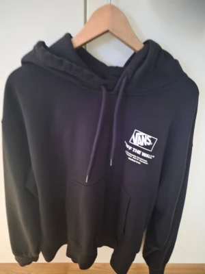 Svart hoodie från Vans med tryck - Säljer en svart hoodie från Vans med vit logga och texten 'Off The Wall' på bröstet. Hoodien har huva med dragsko och en stor magficka framtill. Perfekt för dig som gillar streetwear och vill ha en klassisk look.