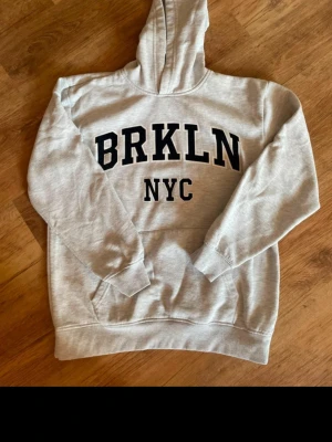Grå hoodie med BRKLN NYC-tryck - Snygg grå hoodie med stor huva och trycket 'BRKLN NYC' i svart på bröstet. Klassisk känguruficka framtill och ribbade muddar vid ärmar och nederkant. Perfekt för en avslappnad streetwear-look. Materialet känns mjukt och bekvämt.