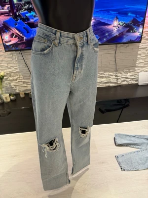 Ljusblå raka jeans med hål - Säljer ett par ljusblå jeans med raka ben och slitna hål vid knäna. Jeansen har hög midja, klassiska fem fickor och är gjorda i denim. Perfekta för en avslappnad och trendig look.