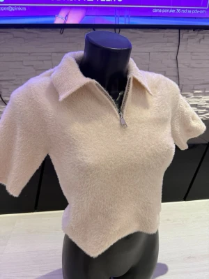 Beige fluffig topp med dragkedja - Supersöt beige topp i fluffigt material med korta ärmar och krage. Dragkedja framtill som kan justeras för olika looks. Perfekt till jeans eller kjol för en trendig och mysig stil.