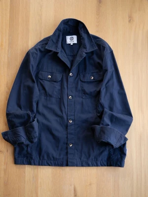 Mörkblå overshirt från Nordam - Stilren mörkblå overshirt från Nordam med klassisk krage och två bröstfickor med knappar. Tillverkad i ett mjukt bomullstyg och har knäppning framtill. Perfekt lager på lager-plagg för en avslappnad look.