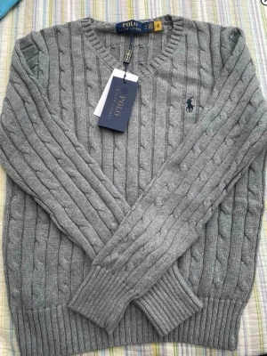 Grå kabelstickad tröja Polo Ralph Lauren - Snygg grå kabelstickad tröja från Polo Ralph Lauren med V-ringning och klassisk broderad logga på bröstet. Tröjan har långa ärmar och ribbade muddar vid ärmslut och nederkant. Perfekt för lager på lager och stilren look.
