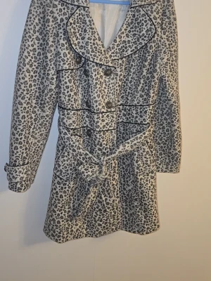Leopardmönstrad trenchcoat med bälte - Snygg trenchcoat med leopardmönster i svart och grått. Jackan har stora slag, knappar framtill och ett matchande knytbälte i midjan. Svarta passpoaler ger extra detaljer. Perfekt för dig som vill sticka ut med din stil.