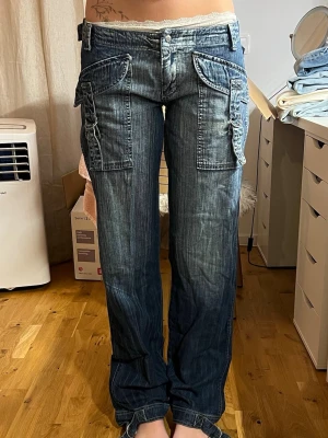 Blå cargobyxor med stora fickor - Säljer ett par blå cargobyxor i denim med bred passform och stora fickor på sidorna. Passar perfekt på mig som är 168 med storlek w27/26, Uppskattar mått midja-37 innerbenslängd-85.