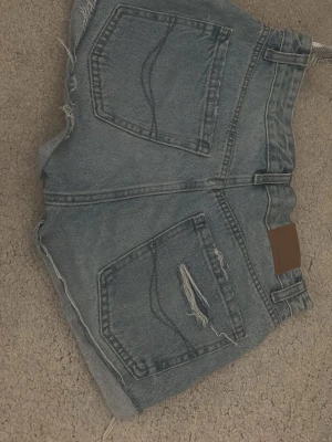 Ljusblå denimshorts med råa kanter - Säljer ett par ljusblå denimshorts med råa, fransiga kanter och slitningar på ena bakfickan. Klassisk femficksmodell med bälteshällor och knappgylf. Perfekta för sommaren och ger en avslappnad vibe.
