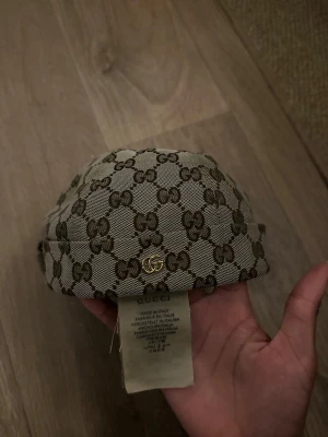 Gucci beige GG-mönstrad kufi - Säljer en beige kufi från Gucci med det klassiska GG-monogrammet i brunt över hela hatten. Tillverkad i canvasliknande material och har en liten guldfärgad GG-detalj på sidan. Perfekt för dig som vill ha en unik accessoar med lyxig känsla.