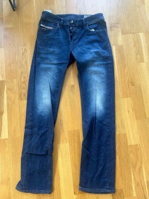 Mörkblå jeans från Diesel - Säljer ett par mörkblå jeans från Diesel med snygg slitning på låren och raka ben. Klassisk femficksmodell med knappgylf och detaljerad logga på myntfickan. Perfekta för dig som gillar en avslappnad men stilren look.