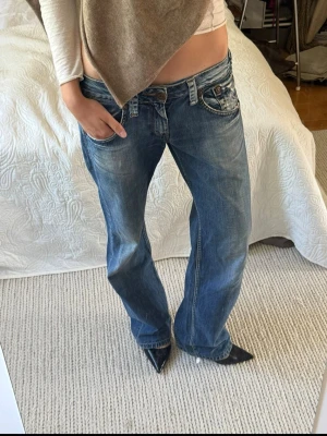 Blå baggy jeans med låg midja - Säljer ett par blå baggy jeans med låg midja och raka ben. Jeansen har slitningar och tvättade detaljer för en avslappnad look. Klassisk femficksmodell med knappgylf och kontrastsömmar. 