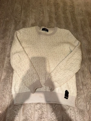 Vit stickad tröja från Fendi - Exklusiv vit stickad tröja från Fendi med diskret FF-mönster över hela plagget. Tröjan har rund halsringning, ribbade muddar och nederkant. Tillverkad i Italien och har en lyxig, mjuk känsla som passar perfekt till din stil.