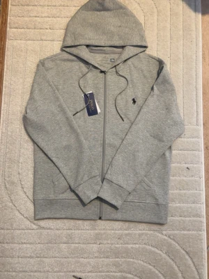 Grå hoodie från Polo Ralph Lauren - Snygg grå hoodie från Polo Ralph Lauren med dragkedja och klassisk huva. Tröjan har dragsko vid huvan, två fickor fram och den ikoniska broderade loggan på bröstet. Perfekt för en chill och stilren look.