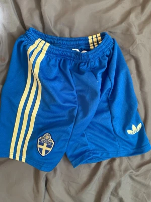 Blå Adidas Sverige shorts - Snygga blå shorts från Adidas med gula ränder på sidorna och Sveriges landslagsemblem framtill. Klassisk Adidas-logga på ena benet. Perfekta för fotboll eller träning, med elastisk midja för skön passform.