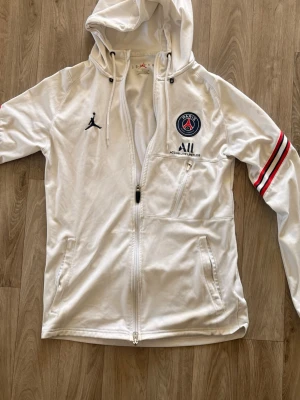 Vit PSG X Jordan huvtröja med dragkedja  - Säljer en vit hoodie med dragkedja och huva, PSG-logga och Jordan-logga på bröstet samt röda och svarta ränder på ärmarna. Två fickor framtill och en extra bröstficka med dragkedja. Perfekt för dig som gillar sportig stil och vill sticka ut.