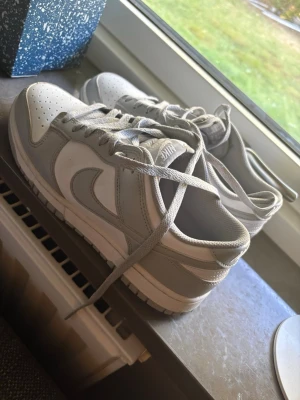 Nike Dunk Low vita/grå sneakers - Nike Dunk Low sneakers i vitt och ljusgrått läder med klassisk Swoosh på sidan. Lågt skaft, perforerad tåbox och matchande vita skosnören. Stilren och tidlös modell som passar till det mesta.