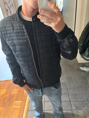 Mörkblå quiltad bomberjacka Jean Paul - Snygg mörkblå quiltad bomberjacka från Jean Paul med dragkedja framtill och två fickor med dragkedja. Jackan har ribbade muddar vid ärmslut och nederkant samt en liten ståkrage med tryckknapp. Perfekt för dig som vill ha en stilren och enkel jacka.