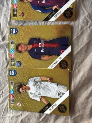 FIFA 365 Guldiga samlarkort - Tre guldiga FIFA 365 samlarkort med fotbollsspelarna Messi, Neymar Jr och Modric. Kortens design har en skimrande guldbakgrund och visar spelarna i sina klubbtröjor från Barcelona, PSG och Real Madrid. Perfekt för dig som gillar fotboll och samlarkort.