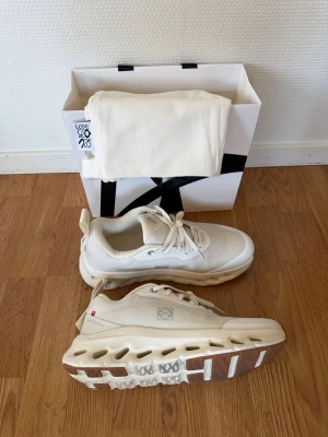 On x Loewe Cloudtilt 2 sneakers - Säljer ett par vita sneakers från On x Loewe med cool sula och vita detaljer. Skorna har mesh och läderpaneler, samt Loewe-logga på sidan och plös. Snygg design med grov yttersula och neutrala färger som passar till det mesta. Storlek 42 men passar till 41