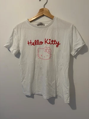 Vit Hello Kitty t-shirt med tryck - Vit t-shirt med rött och rosa Hello Kitty-tryck både fram och bak. På framsidan står det 'Hello Kitty' och en liten Hello Kitty-figur, medan baksidan har en större Hello Kitty och texten 'Happy looks cute on you'. Klassisk passform och rund hals.